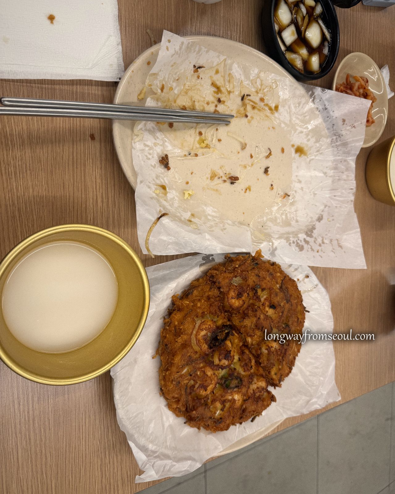 Overhead view of a mung bean pancake on paper beside a brass bowl of makgeolli and side dishes