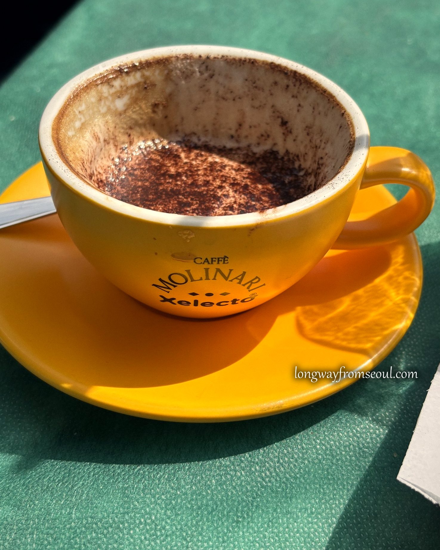 A cappuccino in a yellow Caffè Molinari cup on a green café table in Rome