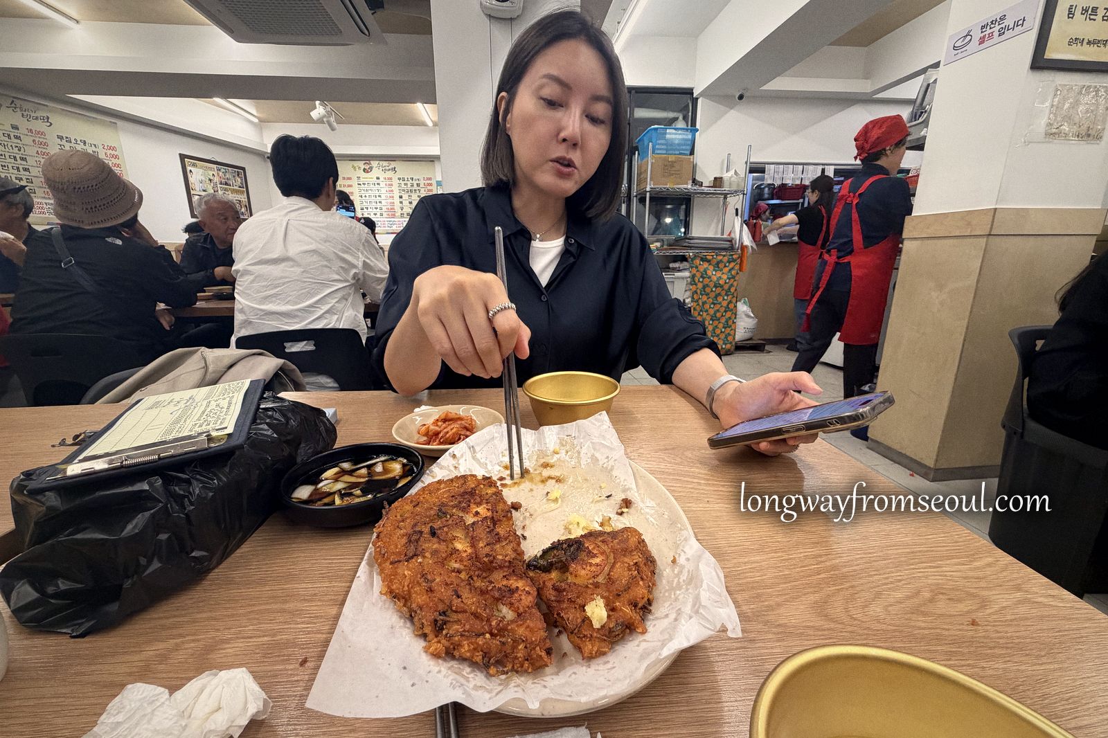 A woman eating at a counter inside Gwangjang Market with stools and other diners behind