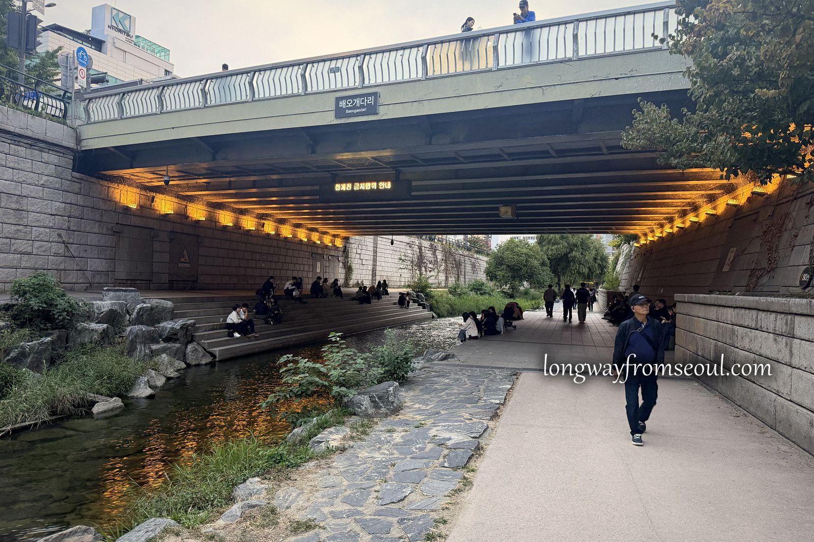 A walkway beside the Cheonggyecheon stream passing under a bridge lit with warm lights