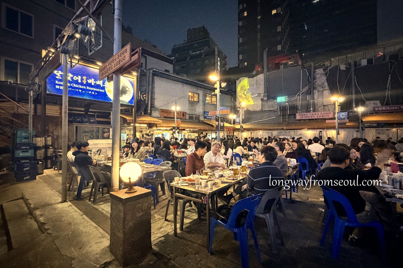 Crowded outdoor tables with blue plastic stools in a grill alley in Euljiro, Seoul, at night