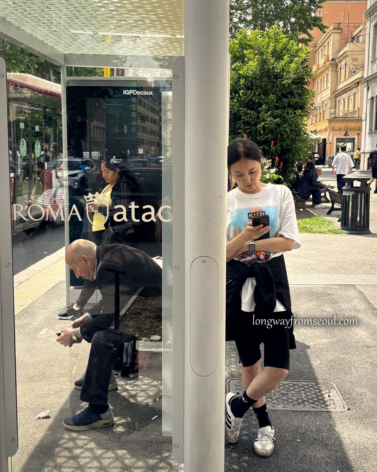 A woman waiting at a ROMA atac bus shelter in Rome, with an older man reflected in the glass