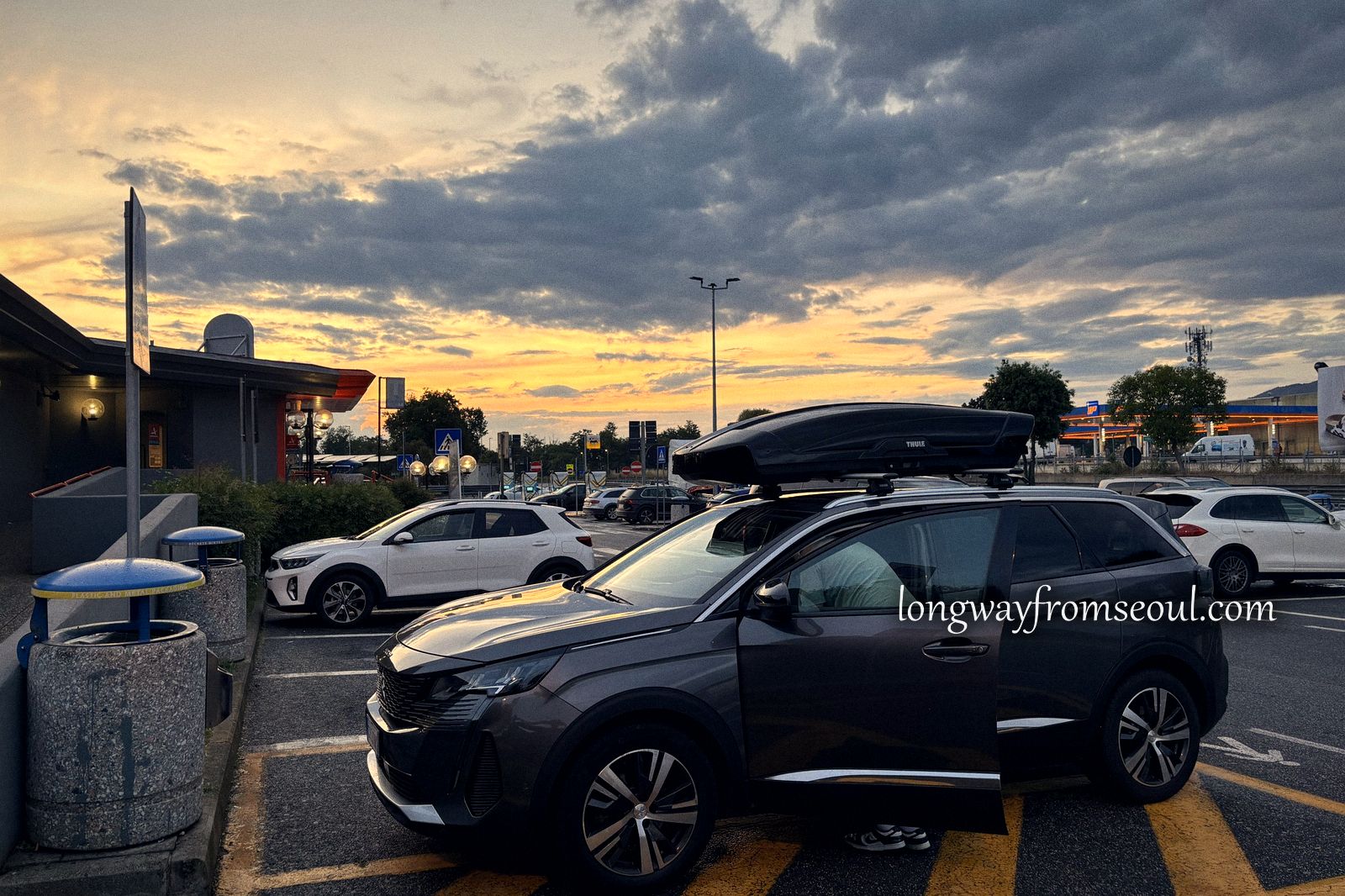 Cars parked at an Italian motorway service station under an orange and grey sunset