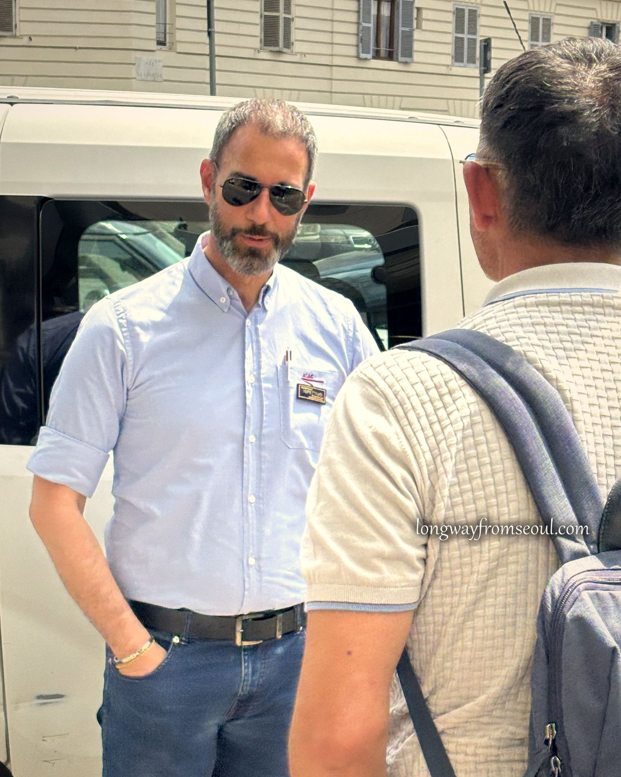 A man in a blue shirt with sunglasses and a name badge standing beside a white minibus in Rome